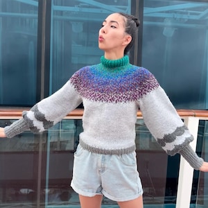 Op de afbeelding: Een handgebreide trui met een hoge groene kraag, die overgaat in blauwe en paarse tinten over de pas. De body is grijs, met grijs-witte gestreepte accenten op de mouwen. Gedragen met lichtblauwe spijkerbroekshorts.