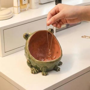 Op de afbeelding: Een groene keramische sieradenschaal in de vorm van een kikker met een perzikkleurig interieur. Een gouden ketting met een hanger wordt boven de schaal gehouden, die ook twee ringen bevat. De schaal heeft gedetailleerde kikkerkenmerken.