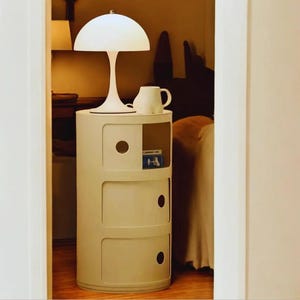 Puede incluir: Una mesita de noche blanca con tres cajones y una abertura circular. Una lámpara blanca con una pantalla en forma de hongo y una taza blanca están encima. La mesa está en un dormitorio.