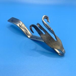 May include: A silver-coloured metal swan-shaped serving utensil, possibly a sugar tong or similar item. The utensil has a curved handle with etched details and a swan head design. The background is a solid light blue.