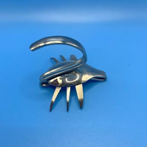 May include: A handcrafted sculpture made from a repurposed fork. The silver-coloured utensil is bent and shaped into an abstract form, with the tines forming legs and the handle curved upwards. The sculpture is set against a solid blue background.