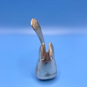 Puede incluir: Un objeto plateado con forma de diente con un elemento en forma de cuchara que se extiende hacia arriba. El objeto tiene dos pequeños agujeros circulares y una forma suave y redondeada. El fondo es azul claro liso.