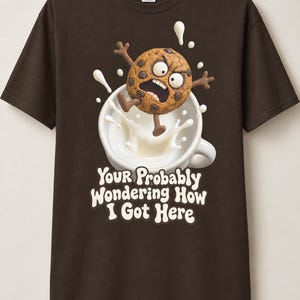 Könnte beinhalten: Ein dunkelbraunes Kurzarm-T-Shirt mit einer Grafik eines schockierten Schokoladenkeks, der in eine weiße Tasse mit Milch springt, inklusive Spritzern. Unter der Tasse steht der Text: "Your Probably Wondering How I Got Here" in weißer Schrift.