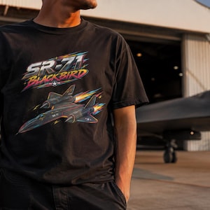 以下が含まれることがあります： SR-71ブラックバード航空機のグラフィックプリントが施された黒いTシャツ。ジェット機の上部にはSR-71 BLACKBIRDの文字がスタイリッシュなフォントで描かれ、機体には青とオレンジのカラフルなモーションストリークと排気炎が表現されています。