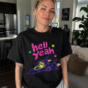 Op de afbeelding: Een zwart t-shirt met een neonroze afbeelding met de tekst "hell yeah" en kleine groene en roze sterretjes. Onder de tekst staat een gestileerde illustratie van een gele kat met een zonnebril die een donkerblauw gevechtsvliegtuig bestuurt.