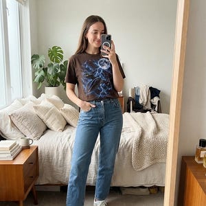 May include: A brown short-sleeved t-shirt featuring a graphic print of a full moon, lightning bolts, and a dark spacecraft. The shirt is paired with blue denim jeans that have a high-waisted fit and a straight-leg cut with a classic button fly.