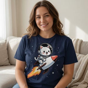 May include: A navy blue short-sleeve t-shirt featuring a graphic print of a white cat wearing a space helmet and suit, riding a white and red rocket ship through space with a bright orange flame trail and small white stars in the background.