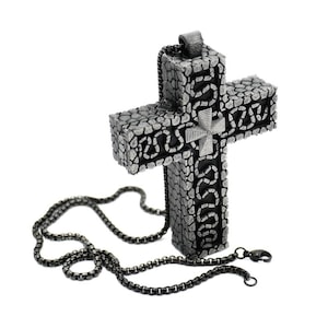 May include: A silver-colored cross pendant with a textured, stone-like appearance. The cross features a detailed design and is suspended from a dark chain. The pendant has a small cross in the center.