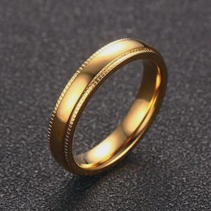 Könnte beinhalten: Ein goldfarbener Ring mit poliertem Finish. Der Ring hat einen detaillierten, strukturierten Rand entlang der Kanten. Der Ring ist vor einem dunklen Hintergrund platziert, der seine reflektierende Oberfläche hervorhebt. Ein klassisches Schmuckstück.