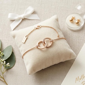 May include: A rose gold bracelet with interlocking circles, adorned with small crystals, rests on a beige pillow. A pair of gold stud earrings is in a clear case. A white bow and a sprig of greenery are also present.