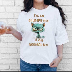 May include: White t-shirt featuring a teal grumpy cat graphic with the text "I'm not GRUMPY this is my NORMAL face" in brown. The cat has an angry expression. The person is wearing blue jeans.