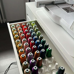 Può includere: Un cassetto pieno di rocchetti di filo colorati, ordinatamente organizzati in un organizer bianco. I colori del filo includono rosso, arancione, giallo, verde, blu, viola e marrone. Sullo sfondo è visibile una macchina da cucire.