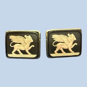 Puede incluir: Un par de gemelos cuadrados con un diseño de león alado dorado sobre fondo negro. Los gemelos tienen un borde dorado y están sobre un fondo azul claro.