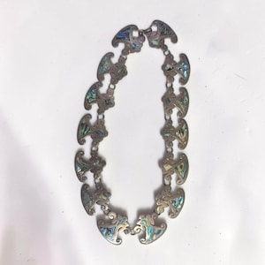 May include: A silver-toned metal link necklace featuring a series of repeating, curved, fan-shaped segments. Each segment is inlaid with iridescent abalone shell pieces displaying shades of green, blue, and purple, connected by small metal rings.