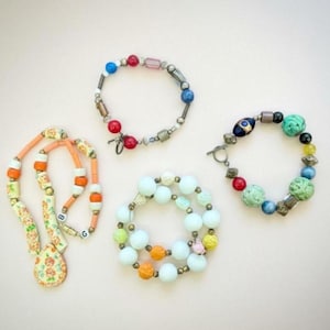 May include: Four pieces of jewellery are displayed on a light background. There is a necklace with orange and white beads and a floral pendant. Three bracelets feature colourful beads in various shapes and sizes, including red, blue, and green.