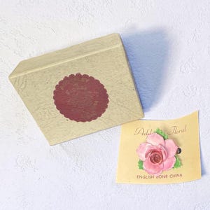 May include: A vintage beige box with a burgundy circular seal. Beside it, a small card displays a pink rose brooch with green leaves. The card reads "Addel Floral" and "English Bone China".
