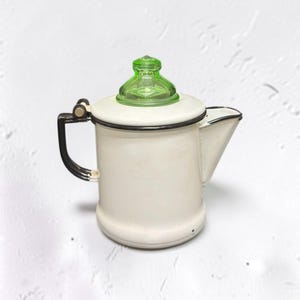 May include: A white enamel coffee percolator featuring a black rim and handle, topped with a translucent, vibrant green glass knob. The cylindrical vessel has a side spout and a hinged lid mechanism, showcasing a classic, utilitarian design.