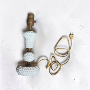 May include: A white hobnail glass lamp base featuring a tiered design with a round base, a spherical middle section, and a flared top. The lamp includes a brass-colored metal socket with a red switch and a long, beige electrical cord with a plug.