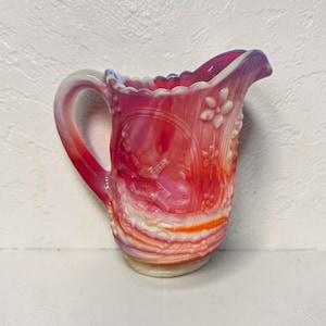 May include: A vintage, ruby-red glass pitcher with a ruffled rim and a handle. The pitcher features embossed floral and geometric patterns. The glass has a swirled, opalescent effect, with shades of red, pink, and white.