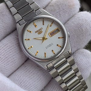 May include: A silver-toned Seiko 5 automatic wristwatch with a white dial. The watch features gold-colored hands and hour markers, and a day-date display. The watch has a stainless steel band.
