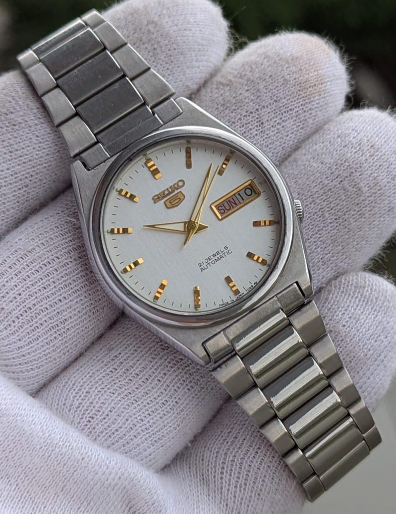 May include: A silver-toned Seiko 5 automatic wristwatch with a white dial. The watch features gold-colored hands and hour markers, and a day-date display. The watch has a stainless steel band.