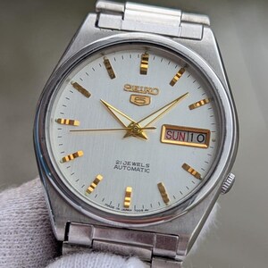 May include: Silver-toned Seiko 5 automatic wristwatch with a white face. The watch features gold-colored hands and hour markers, and a day-date display at the 3 o'clock position. The watch face reads "SEIKO 5", "21 JEWELS AUTOMATIC", and "SUN 10".