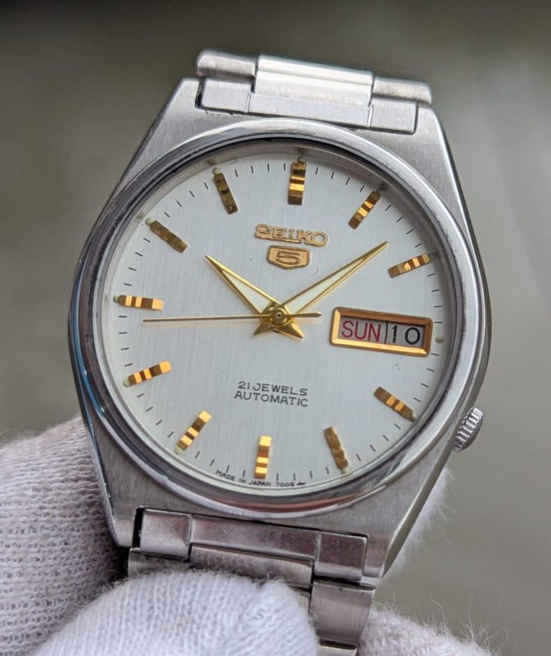 May include: Silver-toned Seiko 5 automatic wristwatch with a white face. The watch features gold-colored hands and hour markers, and a day-date display at the 3 o'clock position. The watch face reads "SEIKO 5", "21 JEWELS AUTOMATIC", and "SUN 10".