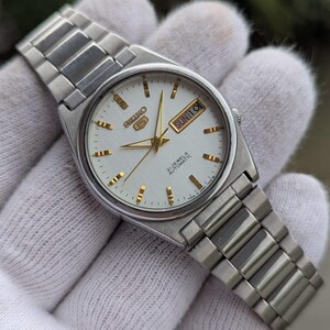 May include: A silver-toned Seiko 5 automatic wristwatch with a white dial. The watch features gold-colored hands and hour markers, a day-date display, and a stainless steel bracelet. The watch face reads "SEIKO 5 21 JEWELS AUTOMATIC".