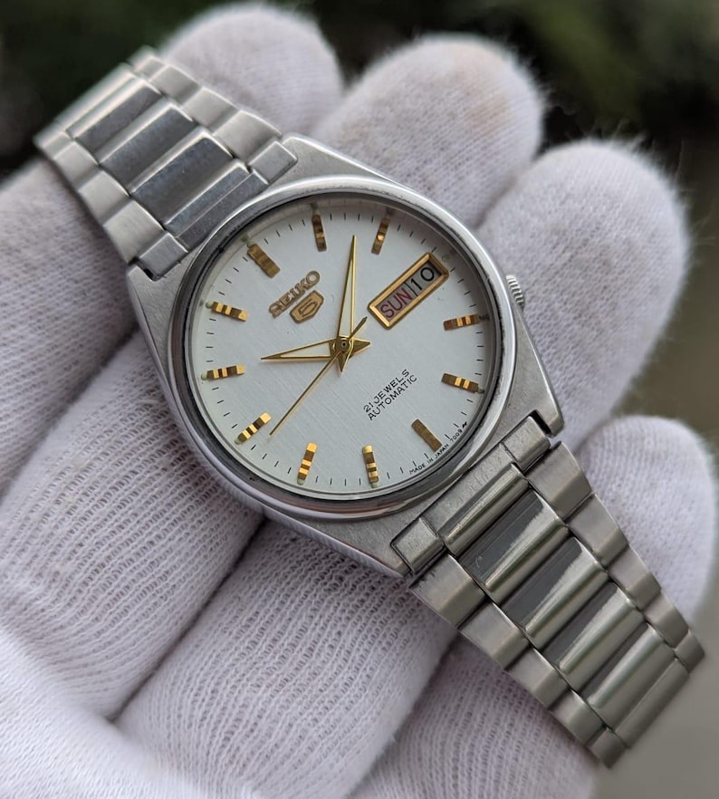 May include: A silver-toned Seiko 5 automatic wristwatch with a white dial. The watch features gold-colored hands and hour markers, a day-date display, and a stainless steel bracelet. The watch face reads "SEIKO 5 21 JEWELS AUTOMATIC".