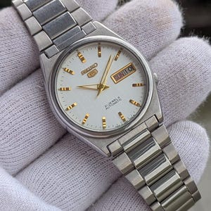 May include: Silver-toned Seiko 5 automatic wristwatch with a white dial. The watch features gold-colored hands and markers, and a day-date display. The watchband is made of silver-toned metal links.