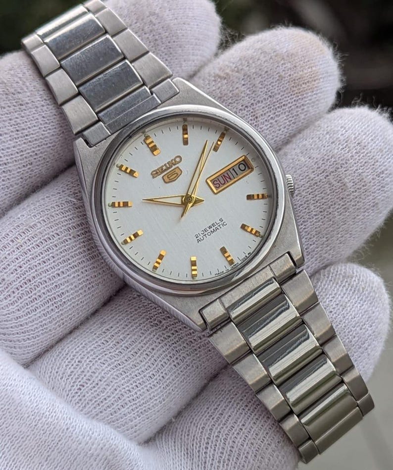 May include: Silver-toned Seiko 5 automatic wristwatch with a white dial. The watch features gold-colored hands and markers, and a day-date display. The watchband is made of silver-toned metal links.