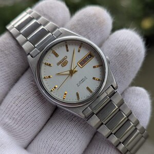 May include: Silver-toned wristwatch with a white face, gold-colored hands and markers, and a stainless steel band. The watch face displays the day and date. The watch is held in a white glove.