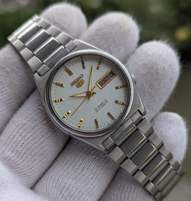 May include: Silver-toned wristwatch with a white face, gold-colored hands and markers, and a stainless steel band. The watch face displays the day and date. The watch is held in a white glove.