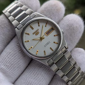 May include: A silver-toned Seiko 5 automatic wristwatch with a white dial and gold-colored hands and markers. The watch face displays the day and date. The watch has a silver metal band.