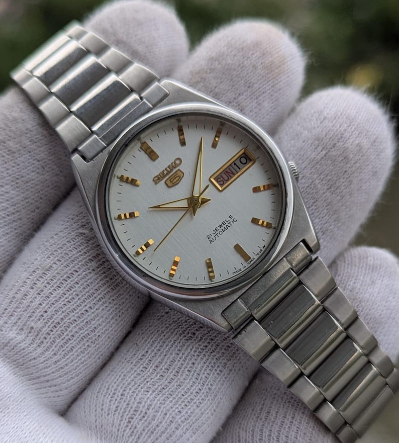 May include: A silver-toned Seiko 5 automatic wristwatch with a white dial and gold-colored hands and markers. The watch face displays the day and date. The watch has a silver metal band.