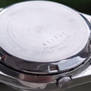 May include: Close-up of the back of a silver Seiko watch. The watch back is stainless steel and features the text "SEIKO", "611795", "WATER RESISTANT JAPAN L", and other markings. The watch is partially visible.