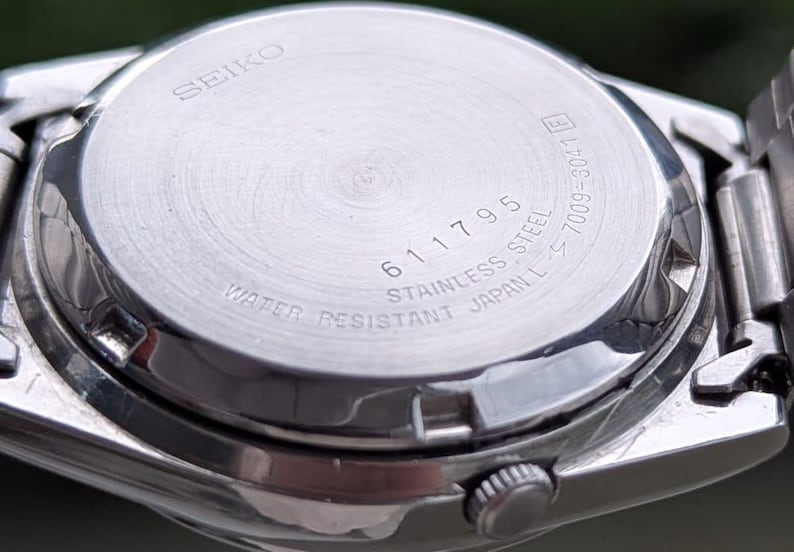 May include: Close-up of the back of a silver Seiko watch. The watch back is stainless steel and features the text "SEIKO", "611795", "WATER RESISTANT JAPAN L", and other markings. The watch is partially visible.