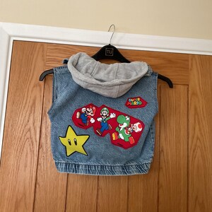 May include: A blue denim sleeveless vest featuring a grey jersey hood. The back displays a large embroidered patch with Mario, Luigi, Yoshi, and Toad, a yellow star patch, and a red Super Mario logo patch. The vest has a ribbed elastic hem.