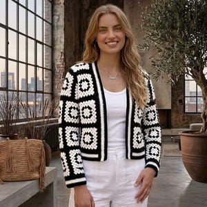 May include: A black and white crochet cardigan with a geometric pattern. The open-front jacket is paired with a white tank top and white pants. A straw bag with tassels sits on a concrete surface.