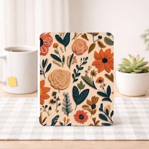 May include: A square coaster with a floral pattern featuring orange, tan, and navy blue flowers and leaves. The coaster is on a light beige background. A white mug with a tea bag and a small succulent plant are in the background.