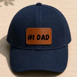 May include: Navy blue baseball cap with a brown leather patch that reads "#1 DAD" in black lettering. The cap has a curved brim and a button on top. A classic and stylish accessory.