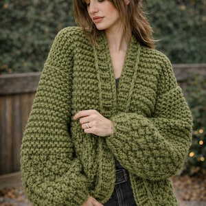 Könnte beinhalten: Ein grob gestrickter Cardigan in gedämpftem Olivgrün. Der offene Pullover hat einen breiten Kragen und dicke, strukturierte Strickärmel. Der Cardigan wird mit einem dunkelgrauen Oberteil und schwarzen Jeans getragen. Der Stil ist lässig und gemütlich.