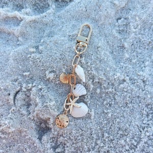 May include: Gold-colored keychain with a lobster clasp and chain, adorned with seashell charms. The charms include a starfish, a sand dollar, and various seashells. The keychain is set against a sandy background.