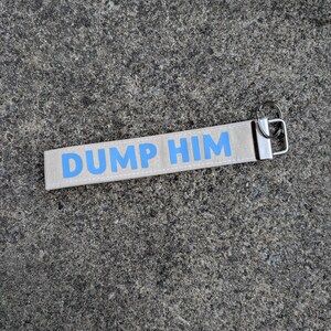May include: A rectangular beige fabric keyring featuring the text DUMP HIM printed in bold light blue capital letters. The fabric strap is attached to a silver-coloured metal hardware clasp and a circular split ring for holding keys.