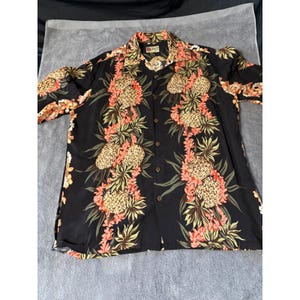 Peut inclure: Chemise hawaïenne noire à imprimé floral. La chemise présente un motif répété d'ananas et de fleurs dans les tons orange, jaune et vert. La chemise à manches courtes est en tissu léger.