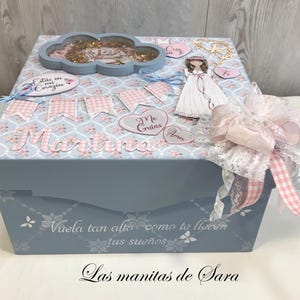 Puede incluir: Una caja decorativa azul claro con un estampado floral y de cuadros. La caja está adornada con un adorno en forma de nube, recortes de papel y un gran lazo rosa. El nombre "Martina" se muestra en la parte delantera. El texto en la caja dice "Vuela tan alto como te lleven tus sueños."
