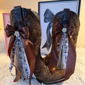 Op de afbeelding: Een paar bruine cowboylaarzen met ingewikkelde bloemenborduursels. Elke laars is versierd met een bruine satijnen strik, een strass-versiering en vloeiende linten in witte en bruine patronen.