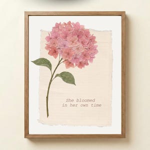 Puede incluir: Una impresión enmarcada con una flor de hortensia rosa prensada con hojas y tallo verdes. La obra de arte está sobre un fondo texturizado blanquecino, con el texto "She bloomed in her own time" en una fuente sencilla.