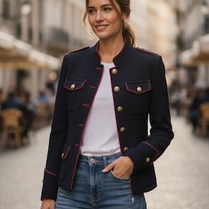 Può includere: Una giacca in stile militare blu navy con colletto alla coreana, profili rossi e bottoni dorati. La giacca presenta due tasche sul petto con patta, maniche lunghe e una vestibilità strutturata, indossata sopra una t-shirt bianca e jeans blu.