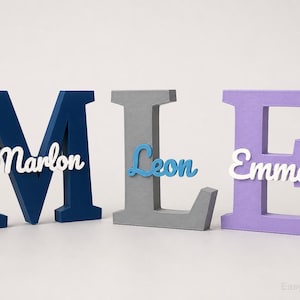 Könnte beinhalten: Drei große Holzbuchstaben in Marineblau, Grau und Lavendel, die "MLE" buchstabieren. Jeder Buchstabe hat einen Namen: "Marlon", "Leon" und "Emma". Die Namen sind in weißer und blauer Schrift.